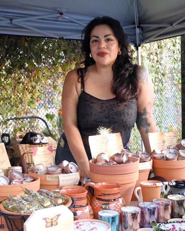 Vendor standing behind a table of handmade pottery and goods under a tent at the market.