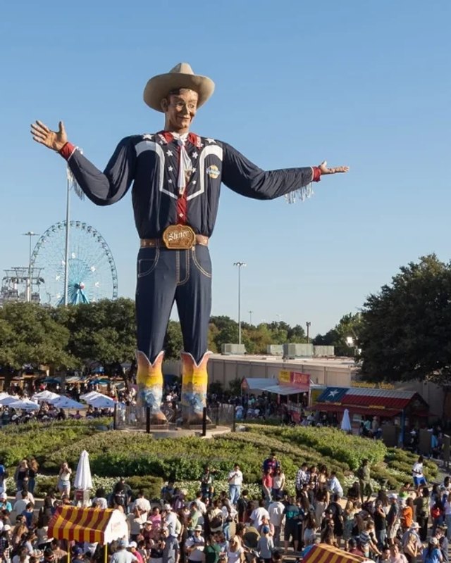 Big Tex statue towering over a crowd at the State Fair of Texas grounds under a blue sky.
