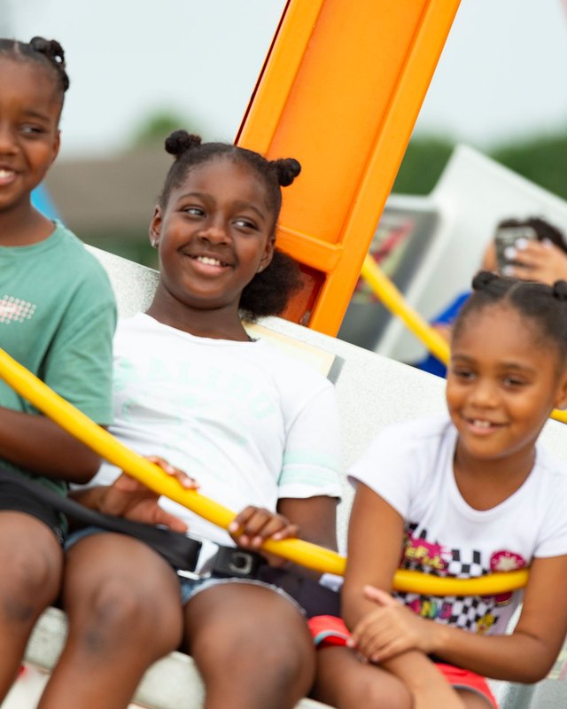 Three smiling girls seated on an amusement ride with yellow safety bars at the 4th Fest; a person in the background holds up a phone.