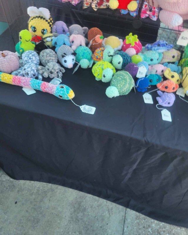 Crocheted plush toys and flowers displayed on a black vendor table at the market.