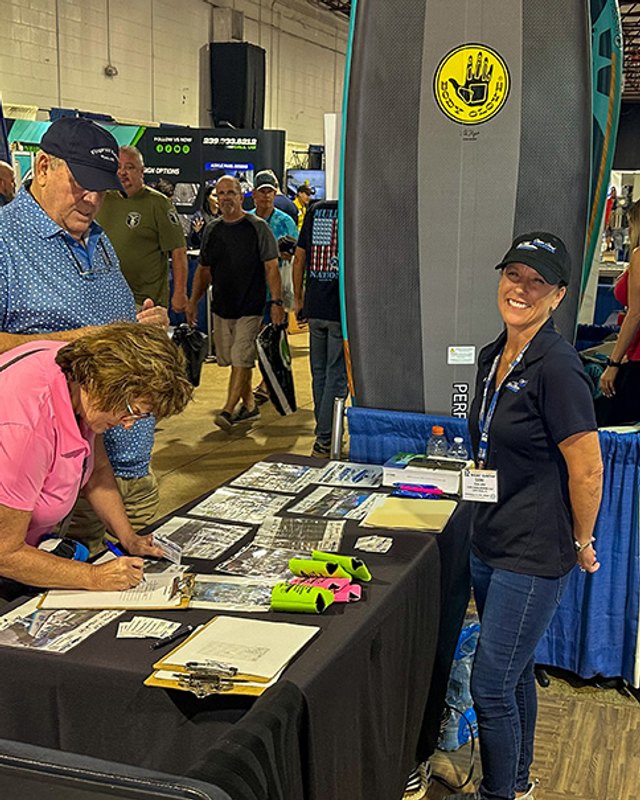 A man signs papers at a boat show exhibitor table while a smiling woman in a black cap stands beside it at the Charlotte County Boat Show.