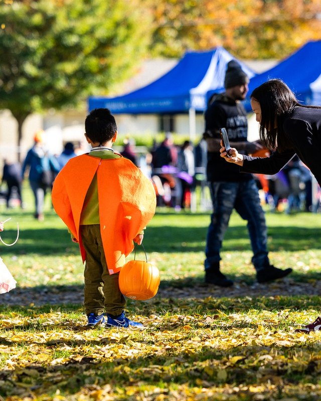 Children in costumes and attendees on a lawn at the Halloween Harvest Festival, with blue event tents behind them.