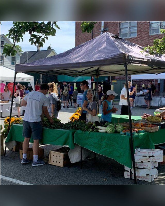 Shoppers gather at a market tent with produce on green-draped tables, with more vendor tents along a street behind them.