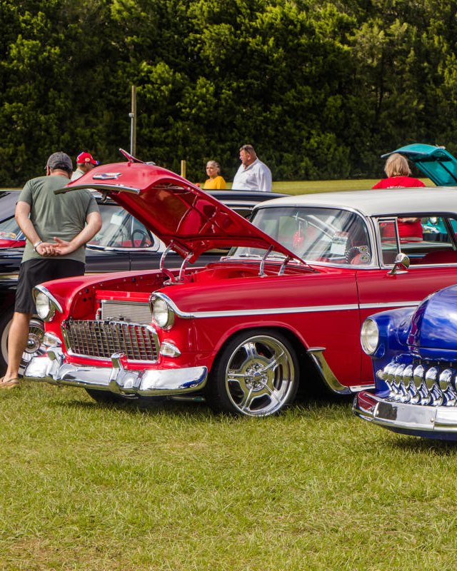 Classic red and blue cars on display at an outdoor car show with spectators around.