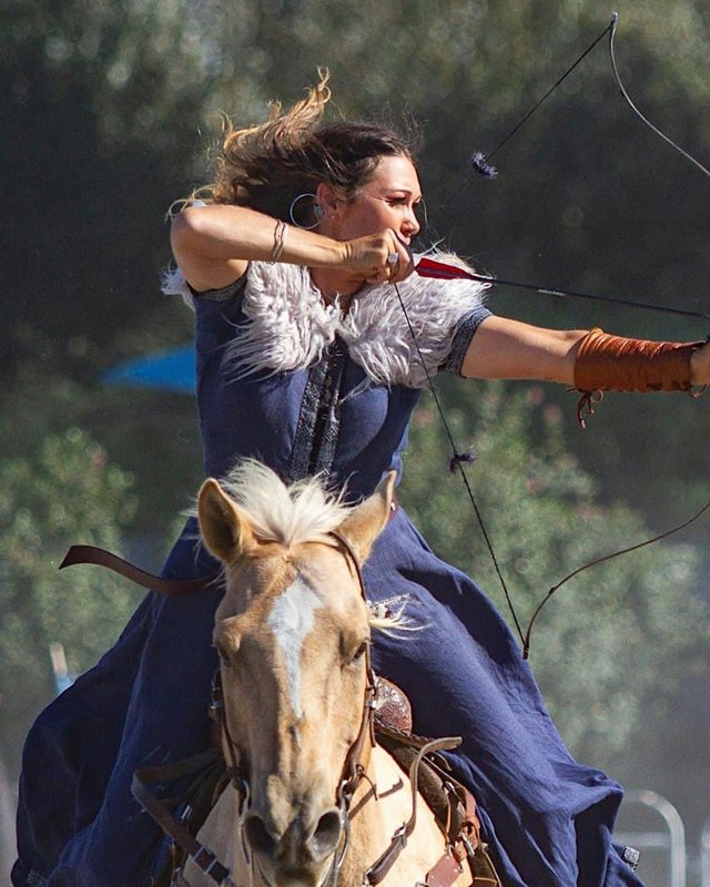 An archer on a horse draws a bow in an arena, with blurred green trees behind.