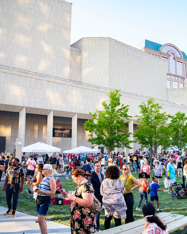 Crowds of visitors walking past white event tents on a sunny lawn outside the Marcus Performing Arts Center