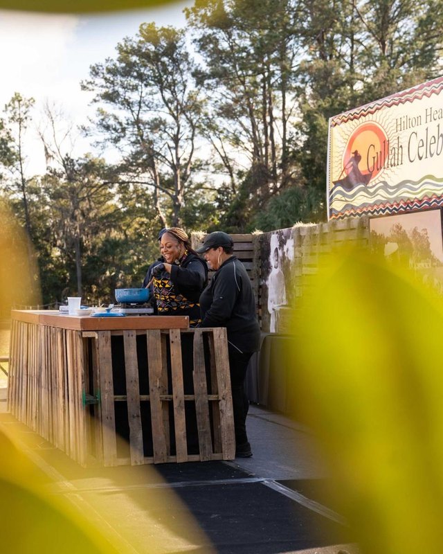 Festival booth with a Gullah Celebration sign, with two vendors working behind a wooden counter outdoors.
