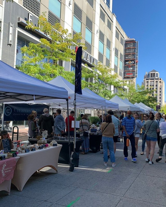 Market booths with tables of goods and shoppers walking under white tents on a downtown Boise street.