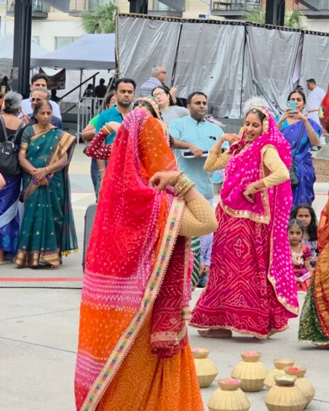 Women in colorful saris dancing among a crowd at an outdoor Puranava festival plaza.