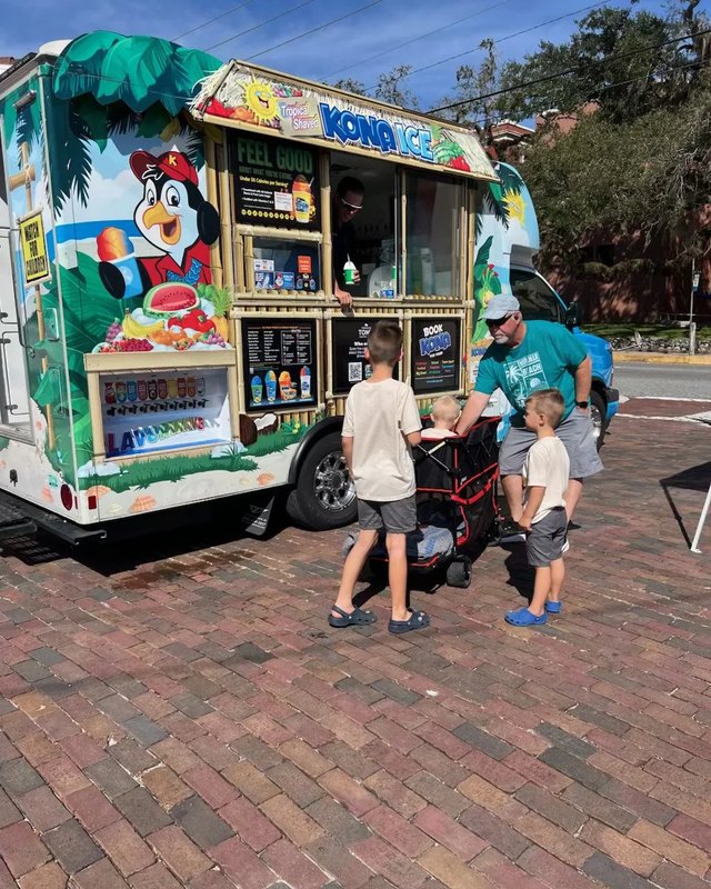 Kona Ice truck with a customer line and a vendor serving kids at a market street.