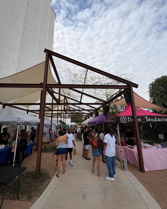 Market stalls under a steel pergola with shoppers walking along a path at the Katy Farmers Market.