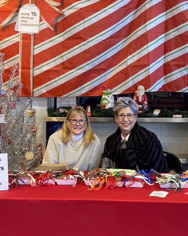 Two women staffing a holiday ornament and ribbon table at a red-draped market booth.