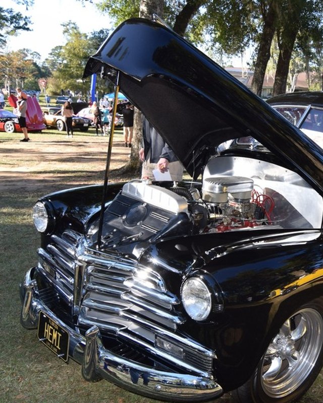 Black vintage sedan with its hood open on display at the Celebration Car Show, with spectators on the grass behind.