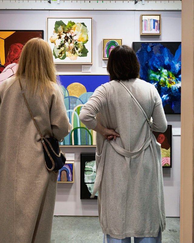 Two visitors in long coats viewing colorful paintings on the walls of an art fair booth.