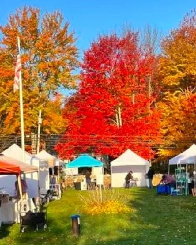 Outdoor market with white and colored vendor tents and shoppers under autumn foliage