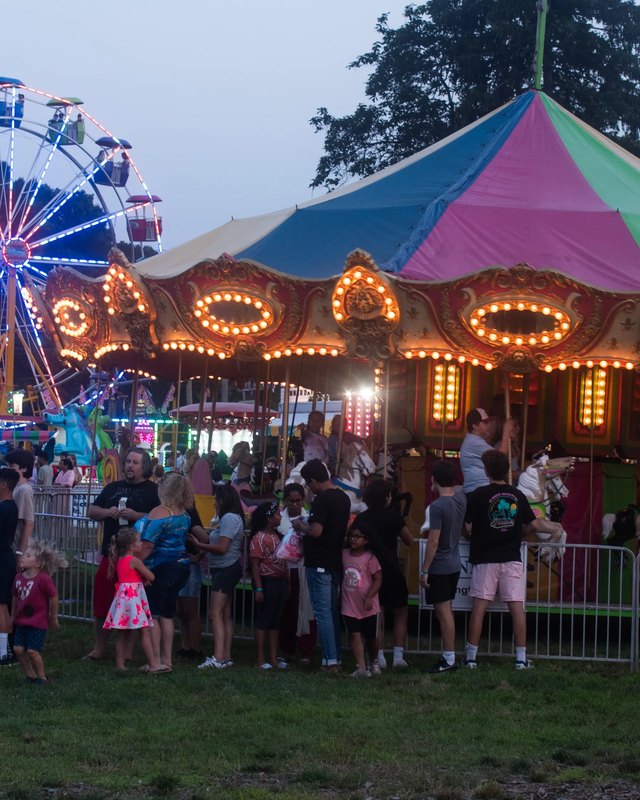 Evening crowd at Vernon's Summer Days Carnival by the Ferris wheel and a lit carousel tent.