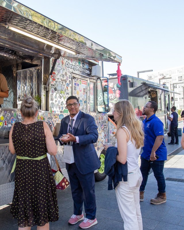 Visitors chatting beside a food truck covered in stickers at uCity Square, with a man in a navy suit at center.