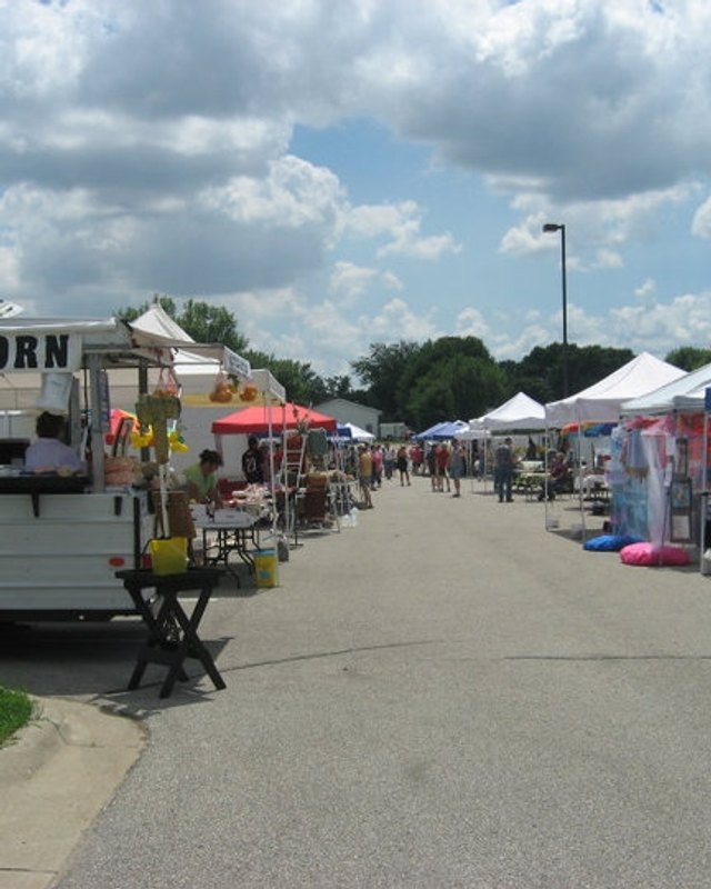Farmers market street with white and red canopy booths and a food trailer with a partly visible 'RN' sign, shoppers browsing under a cloudy sky.