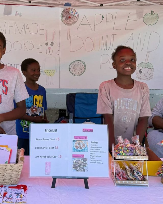 Kids standing behind a craft and treat sales table with baskets and a handmade sign at a community fair.