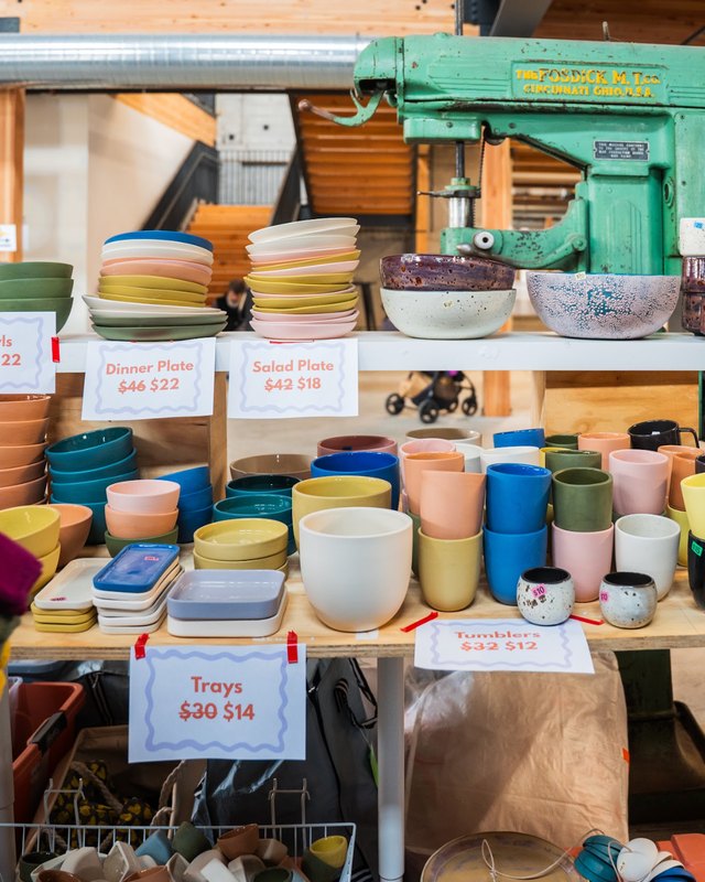 Market booth stacked with colorful ceramic mugs, bowls and plates, with handwritten price tags and a green vintage machine behind.