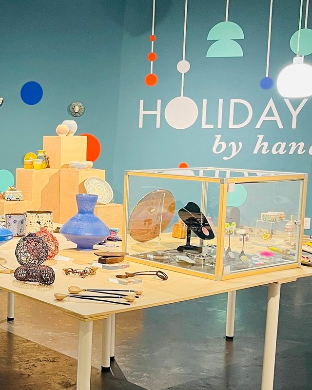 Craft goods on a display table, including ceramic vases and jewelry in a glass case, under a HOLIDAY by hand wall sign.