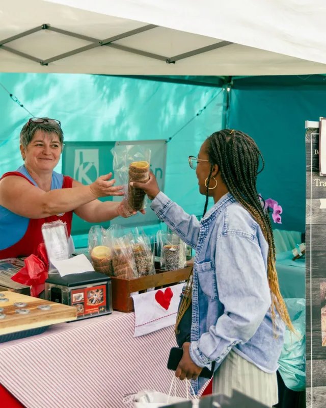 Vendor at a market booth handing a jar of goods to a customer under a white tent