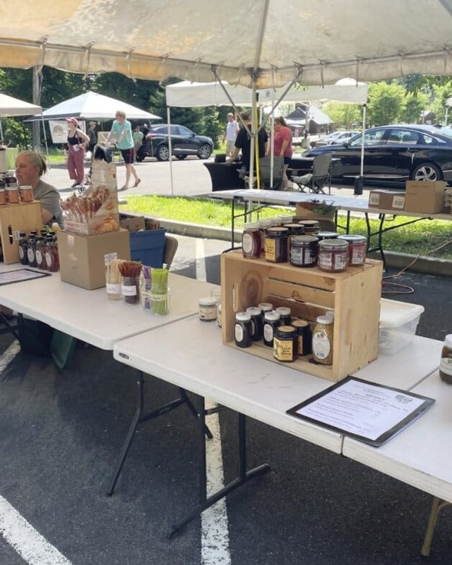 Farm market tables with jars of preserves under a white tent in a parking lot, with shoppers in the background.