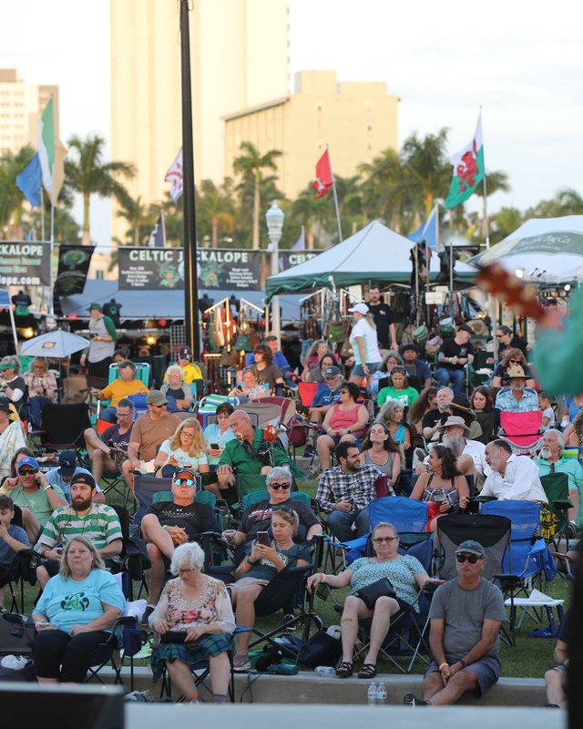 Outdoor crowd seated in folding chairs before white booth tents and flags at the Caloosahatchee Celtic Festival grounds