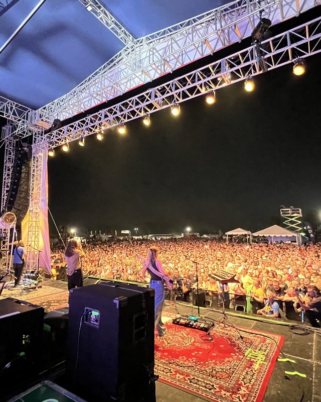Wide stage with lighting truss and a large crowd facing it at night during a festival.