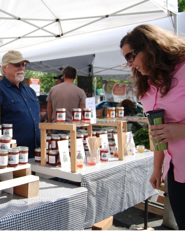 Shopper and vendor at a market booth stocked with jars of preserves under a white canopy.