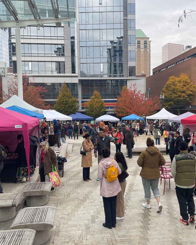 Crowd walking past pink and white vendor tents in a plaza at Director Park during a fall event.