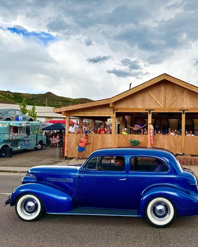 Blue classic car parked outside a wooden outpost building with a crowd and food truck nearby