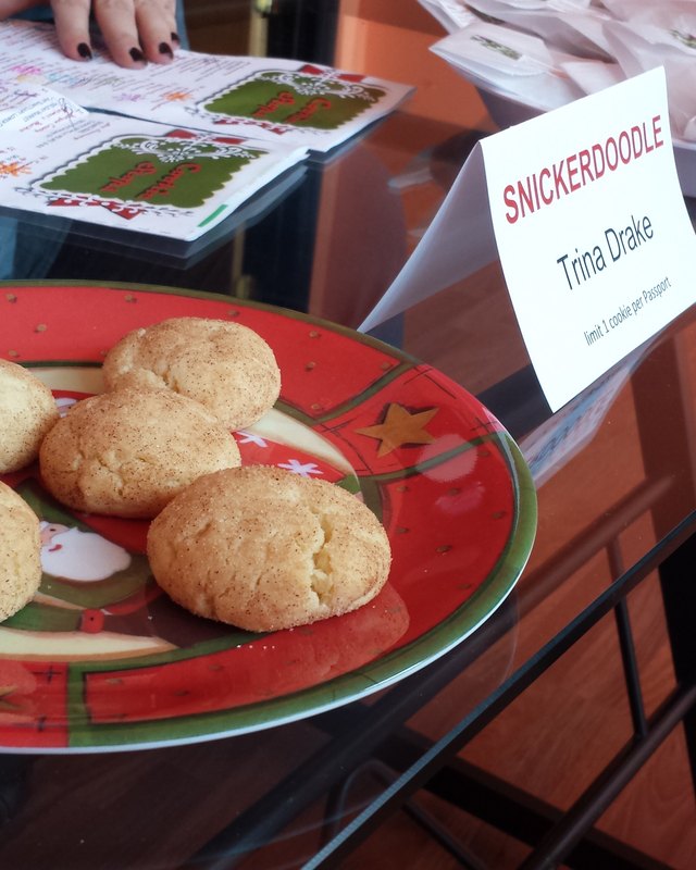 Snickerdoodle cookies on a red holiday plate beside a vendor sign reading 'Snickerdoodle, Trina Drake' at a cookie stroll table.
