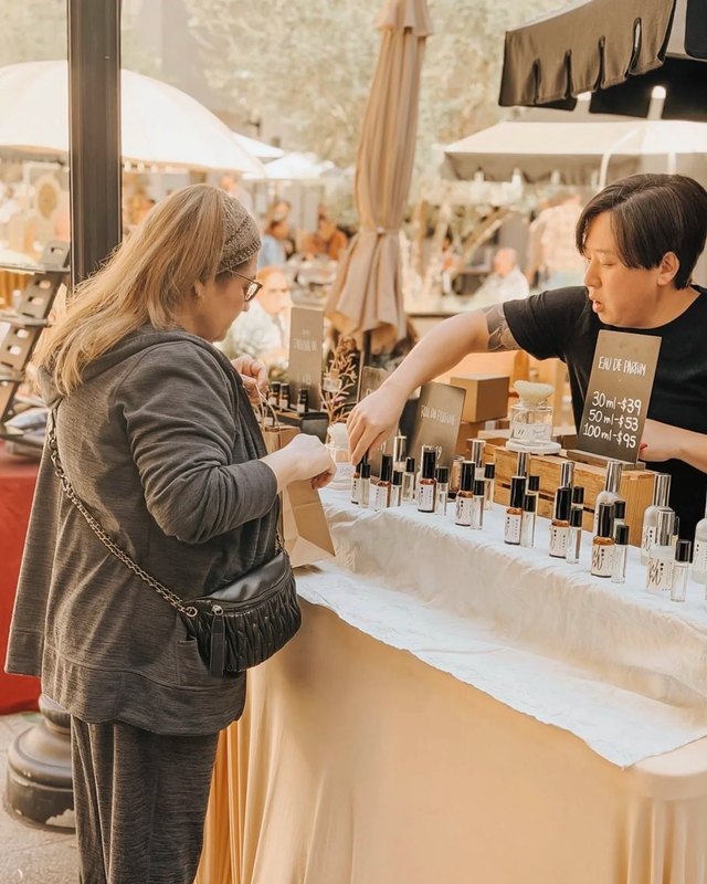 A shopper looks at a vendor's table of small bottled products, with a vendor behind it, at an outdoor market