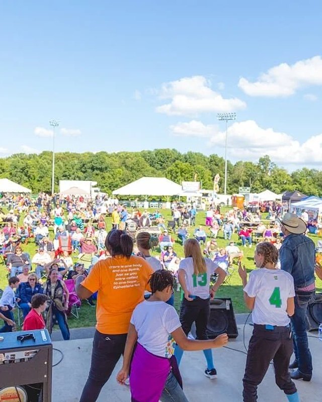 Festival crowd on a grassy field with white tents and vendor booths under a blue sky