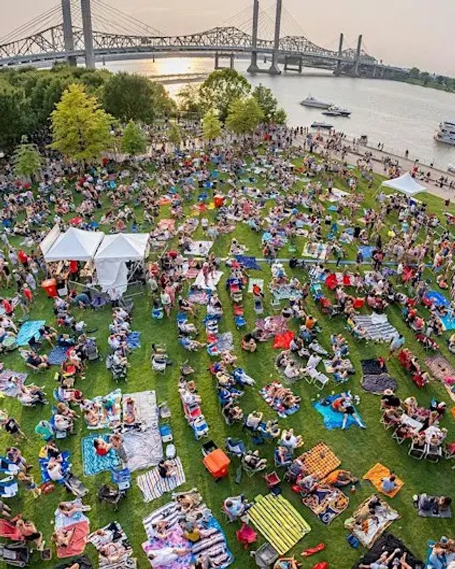 Aerial view of a large crowd on a riverside lawn with white tents and bridges over the river.