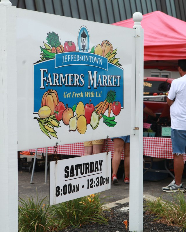 Jeffersontown Farmers Market sign with red tents and shoppers behind it on Saturday mornings