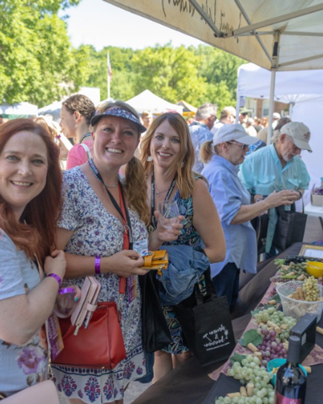 Festival attendees smile beside a vendor table with grapes and wine bottles at an outdoor wine festival.