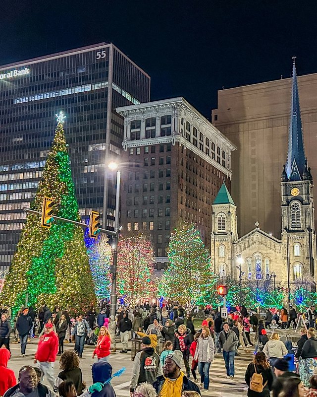 Crowd gathered around a lit holiday tree in front of a church and downtown buildings at night