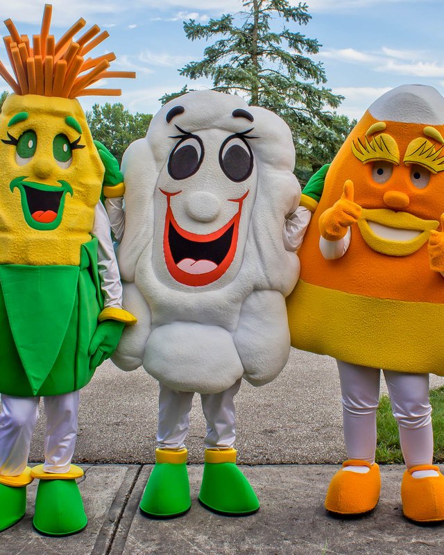 Three costumed mascots, one corn, one popcorn and one candy corn, posing side by side on a sidewalk at a past North Ridgeville Corn Festival.