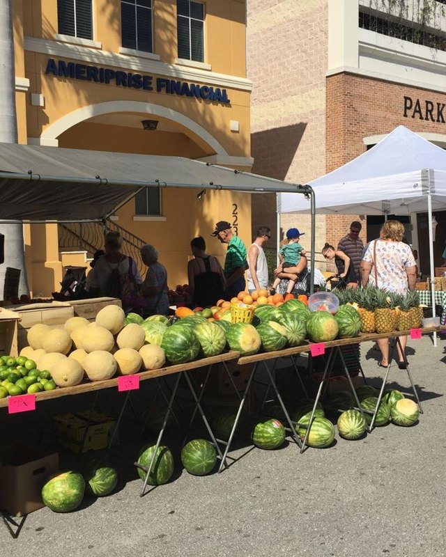 Produce stand with melons, limes and watermelons under a tent, with shoppers in front of a yellow building with an Ameriprise Financial sign in downtown Punta Gorda.