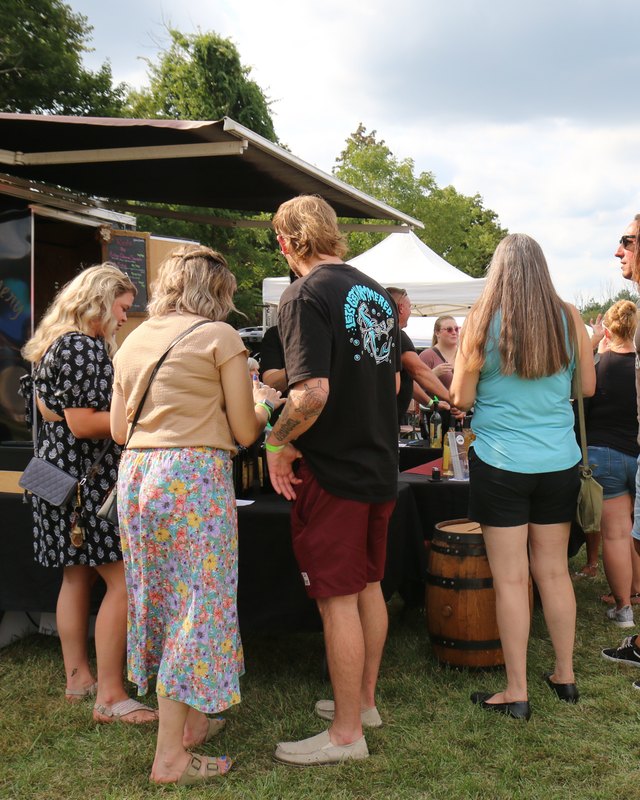 Visitors at a wine tasting table with bottles and a wooden barrel at an outdoor Wines On The Hill booth on a partly cloudy day.