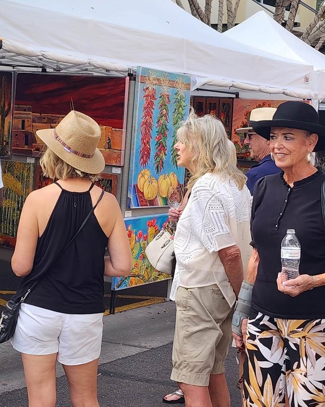 Shoppers in hats looking at colorful paintings on display at an outdoor art festival booth.