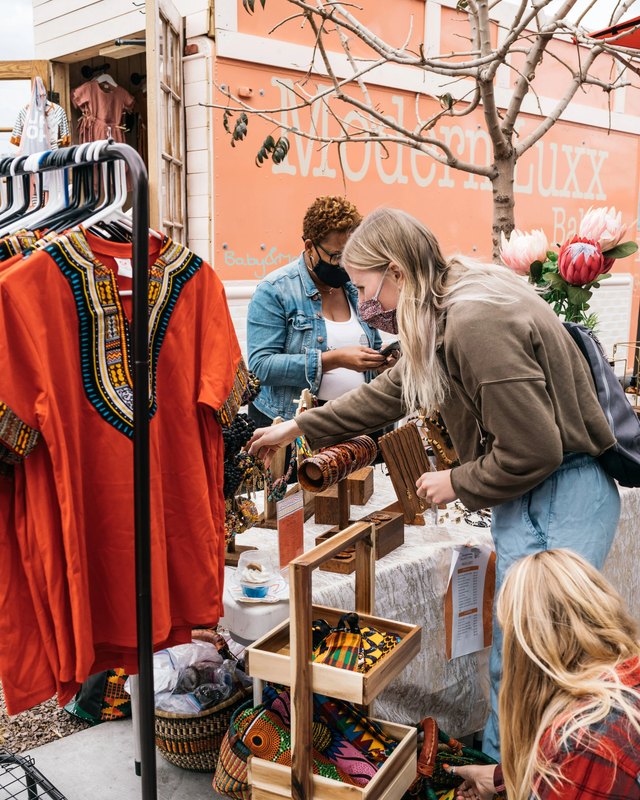 Shoppers browsing an outdoor market stall with African-inspired clothing, jewelry and woven goods on display.