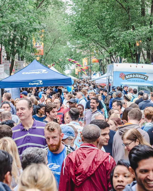 Dense crowd walking along a Philadelphia street lined with blue vendor tents and a Ben & Jerry's truck during a street fair.
