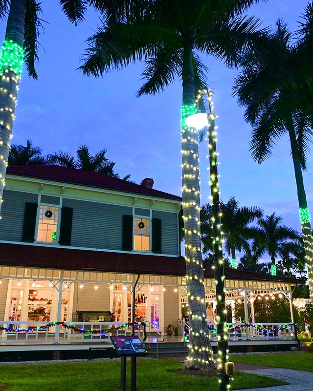 Historic Edison and Ford estate home wrapped in holiday lights with green-lit palm trees at dusk.