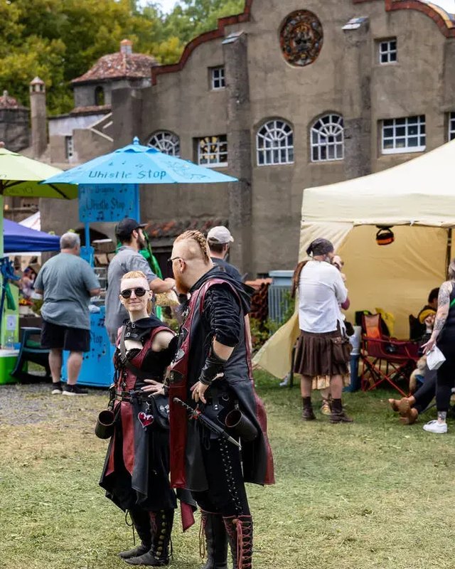 Costumed attendees stand near vendor tents and a stone building with arched windows at the Bucks County Renaissance Faire