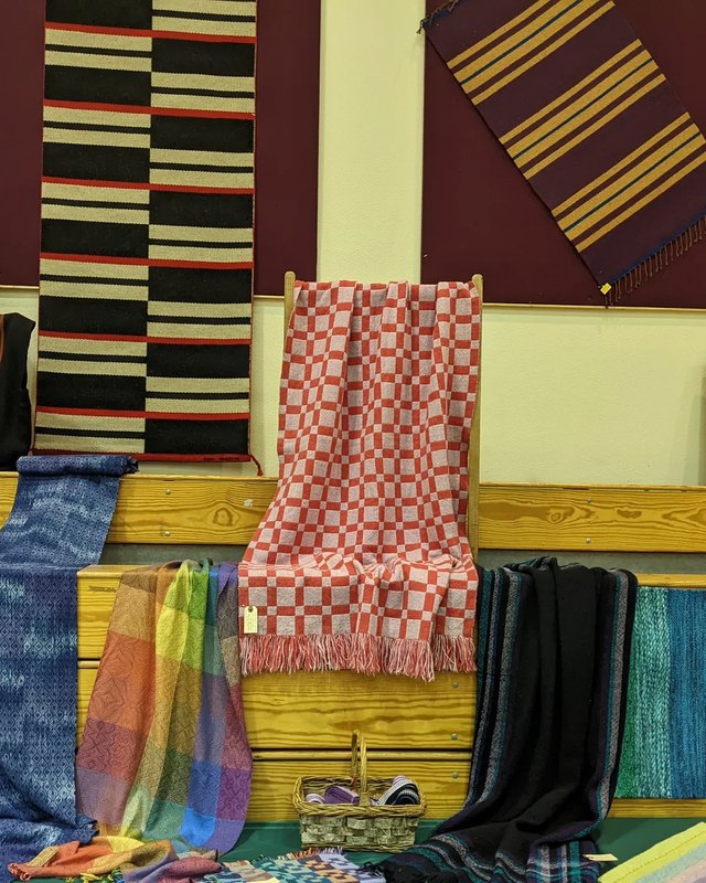 Handwoven blankets and scarves hung for display in a hall, with a basket on a table