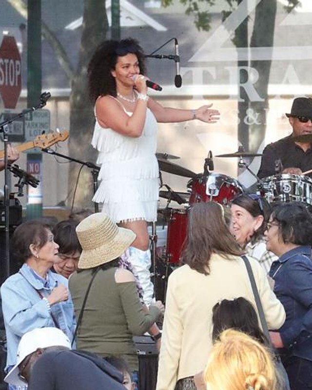 Female singer performing at a microphone on stage beside a drummer, with a crowd in front at a Tracy block party.