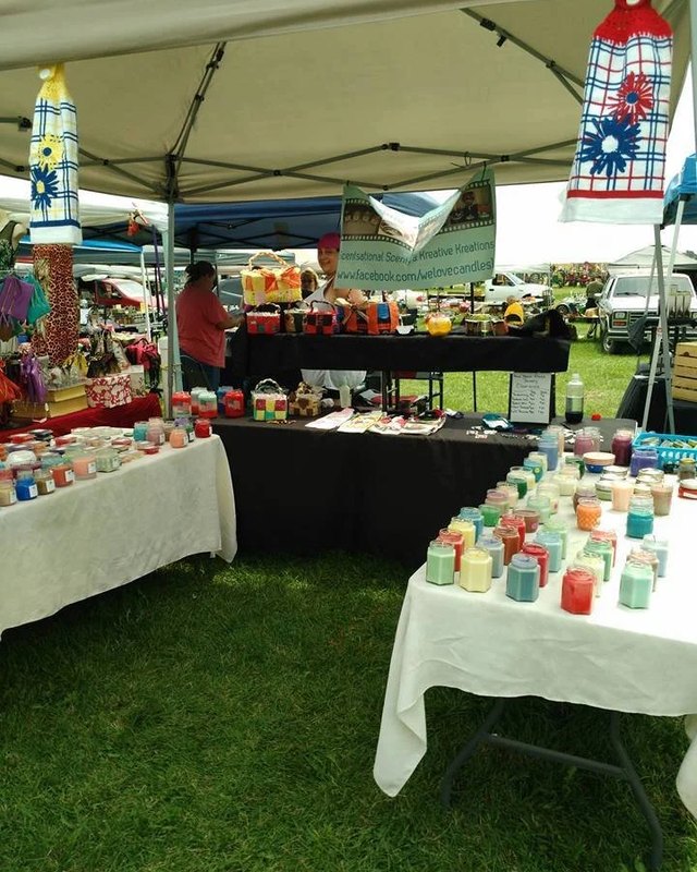 Craft show booth with rows of colored glass jars on white-covered tables under a white canopy, with a patterned banner hanging above and a dark table behind.